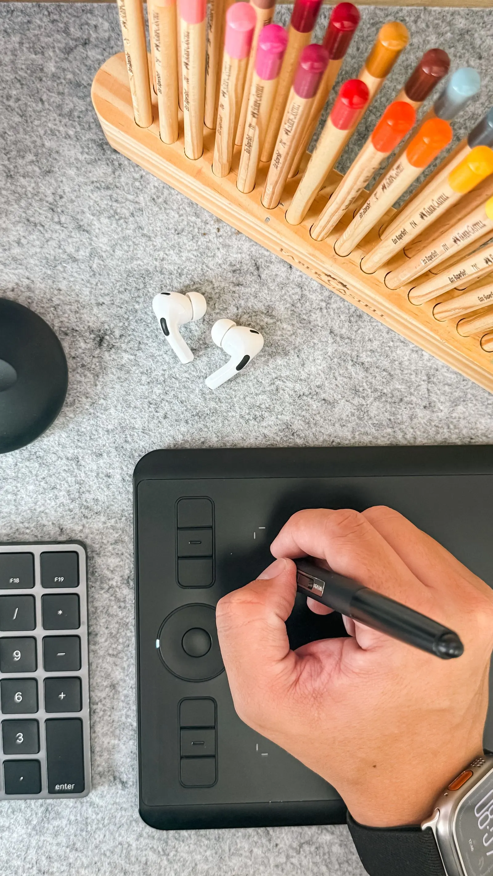 A hand draws on a graphics tablet beside a fan of coloured pencils.