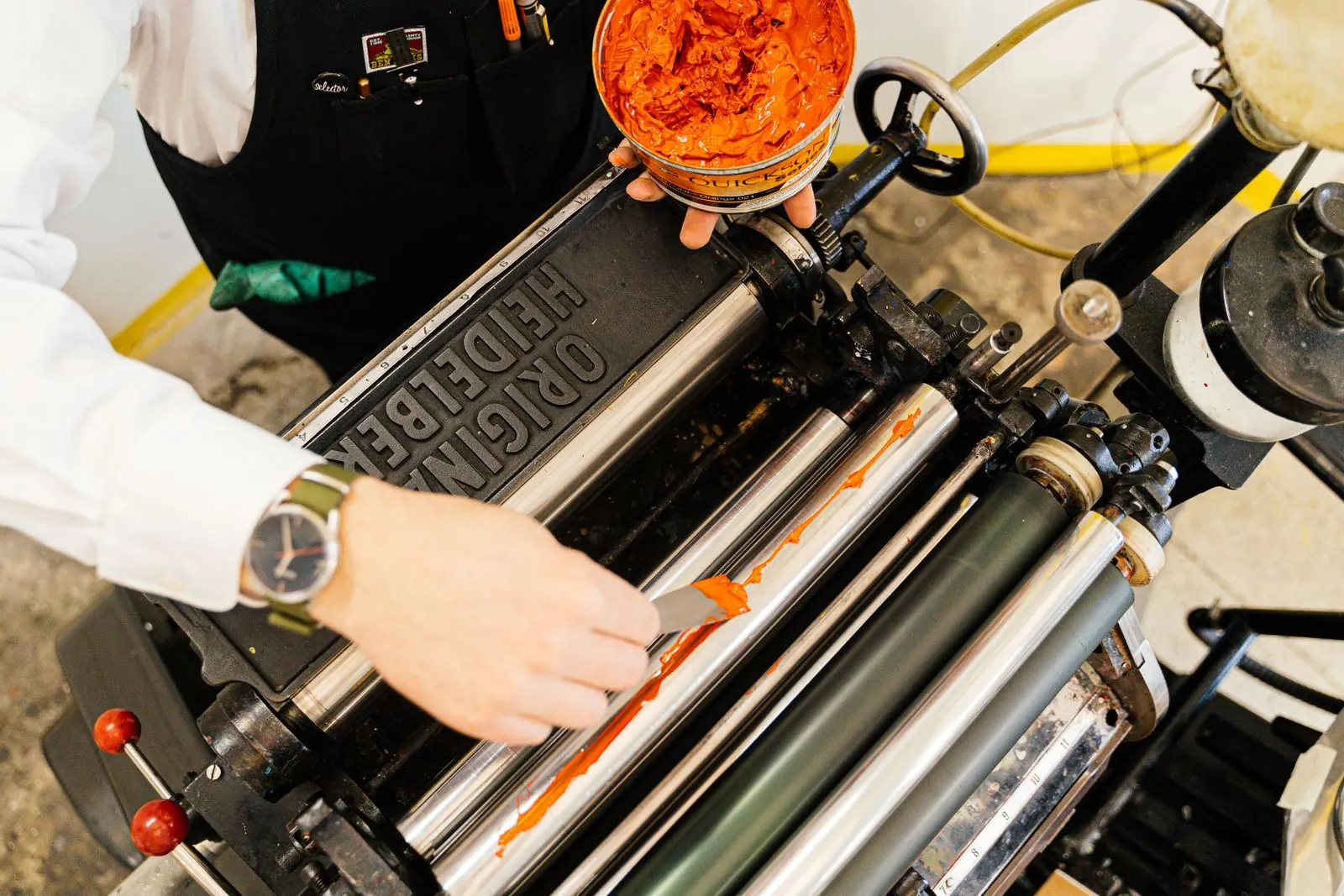 A printmaker applies bright orange ink to the rollers of a letterpress.