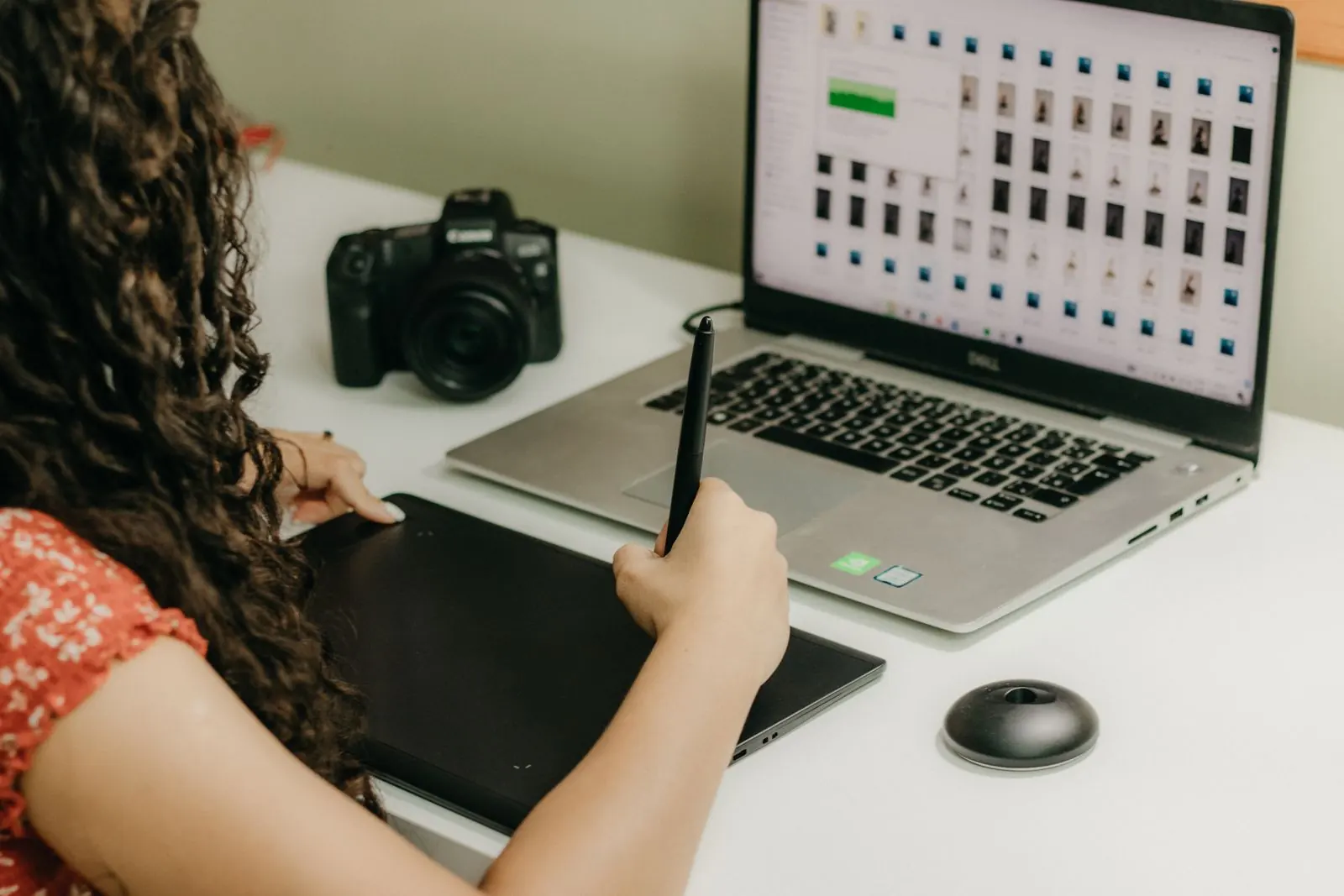 A designer uses a graphics tablet beside a laptop showing a grid of visual designs.