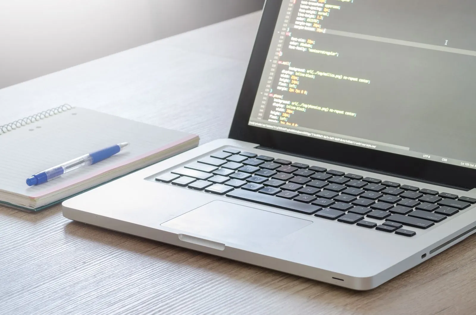 A laptop shows source code beside handwritten notes and a blue pen on a white desk.