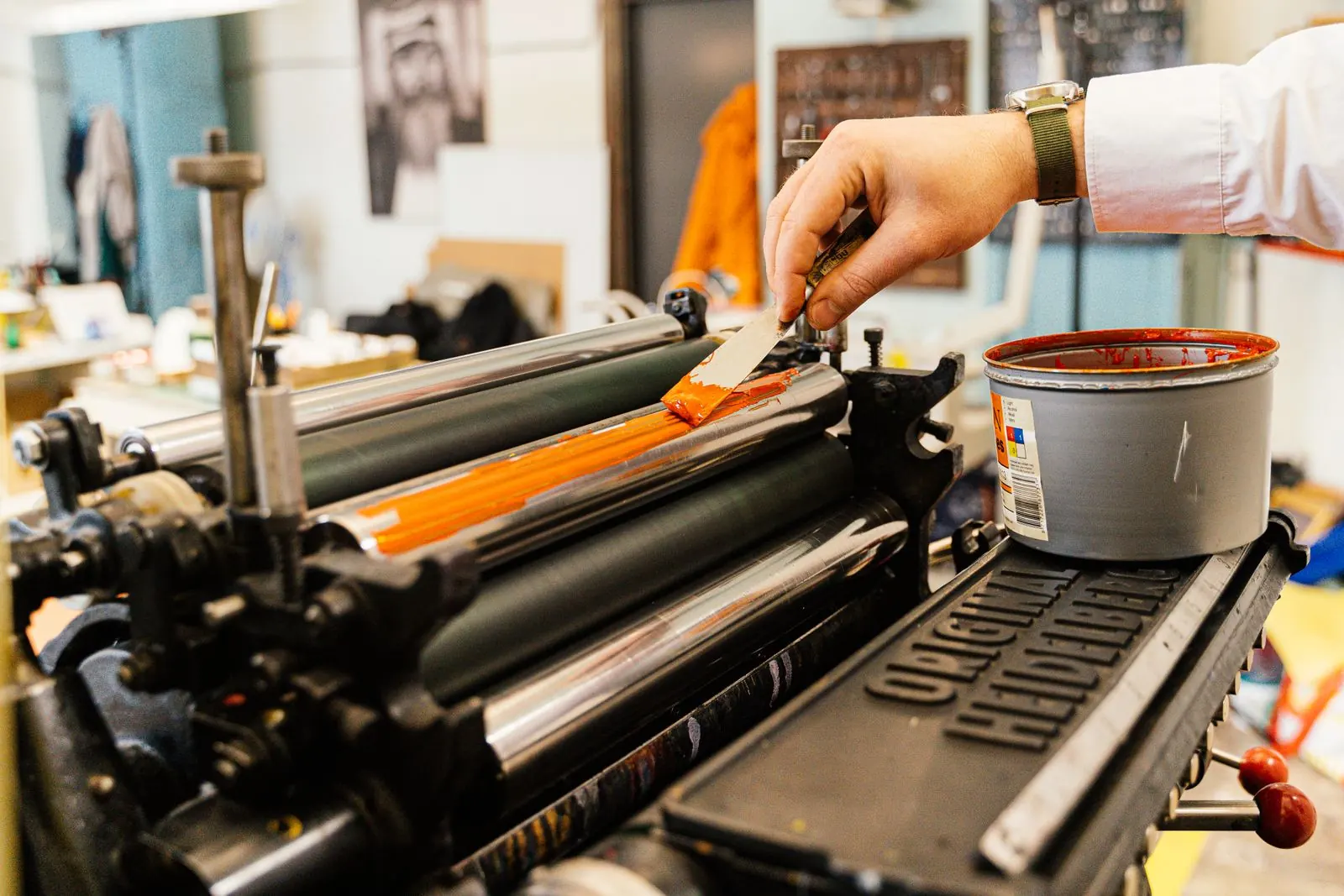 A gloved hand spreads orange ink onto rollers inside a printing press.