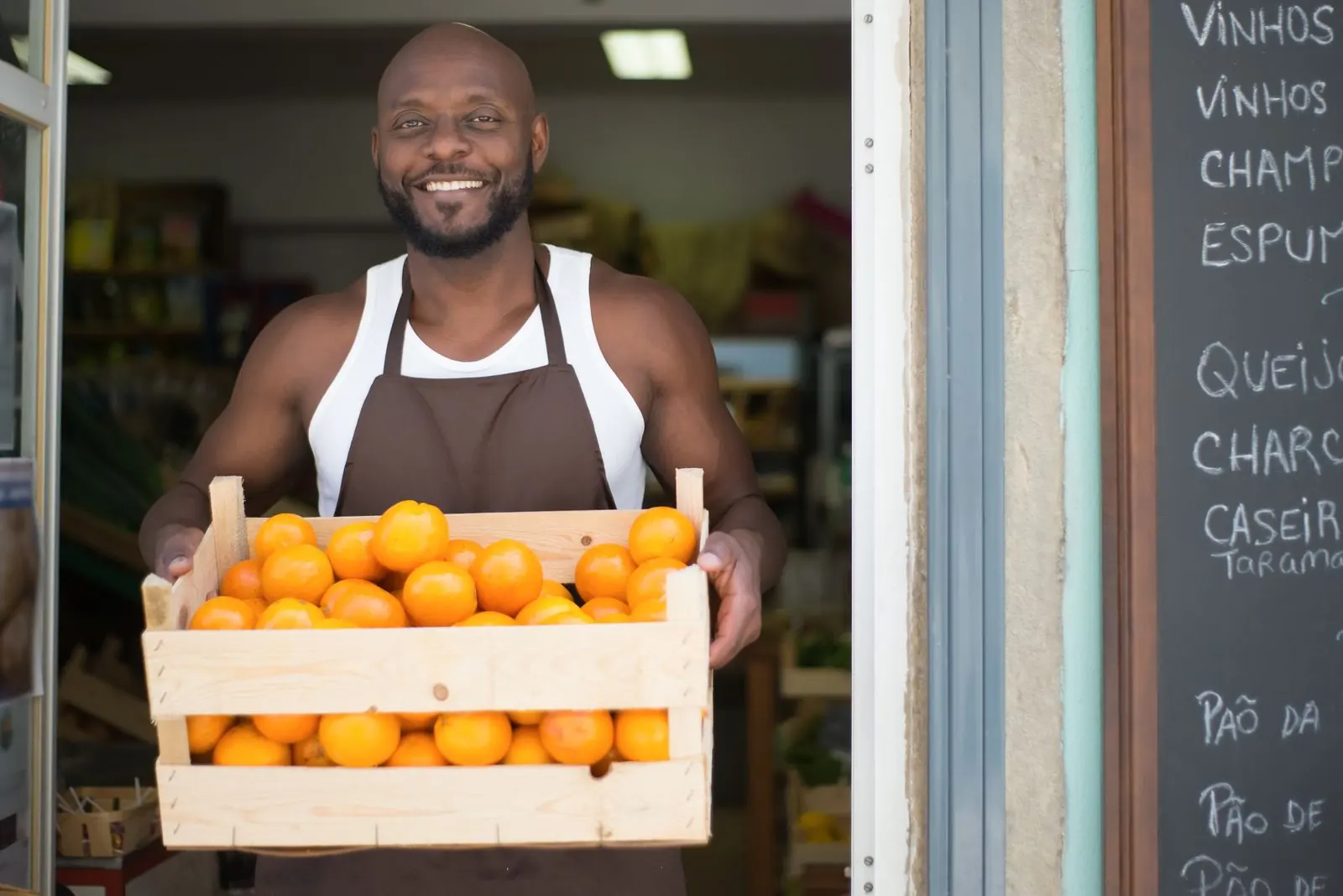 A smiling person holds a crate of oranges in the doorway of a small café.