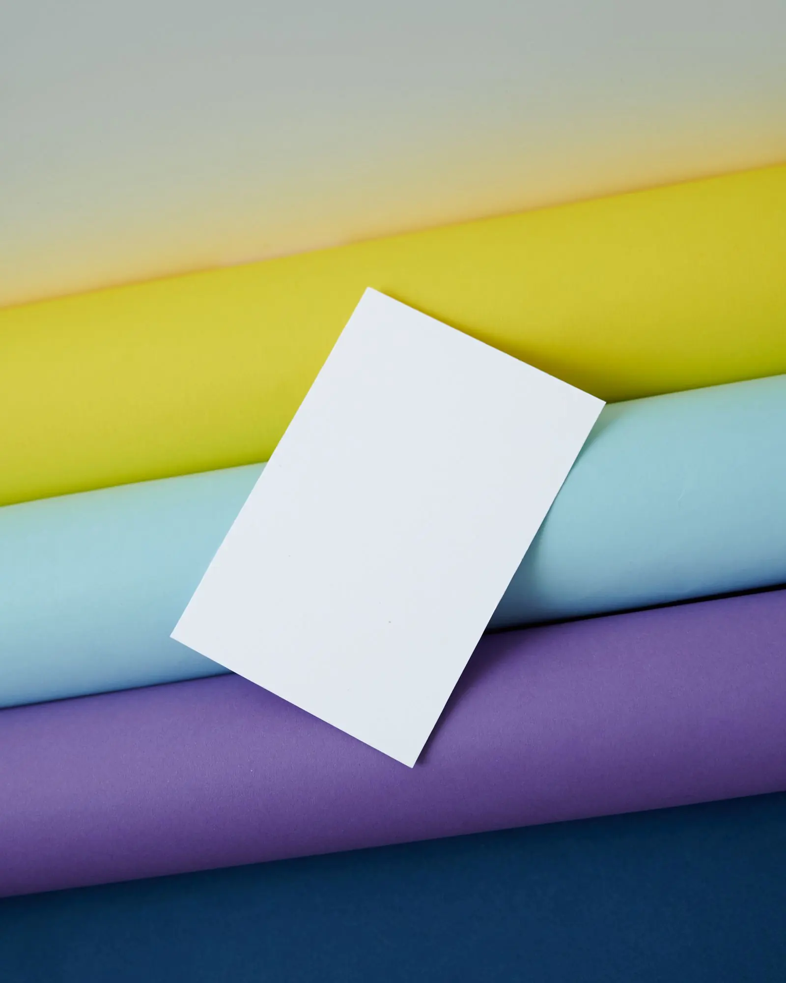 A blank white card rests diagonally across yellow, green, blue and purple rolled paper.