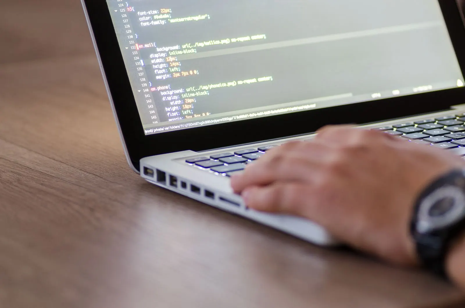 Hands work at a laptop displaying code on a wooden tabletop.